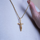 A gold crucifix pendant necklace worn on a person's neck, shown against a white t-shirt with a hand holding the chain near the collarbone.