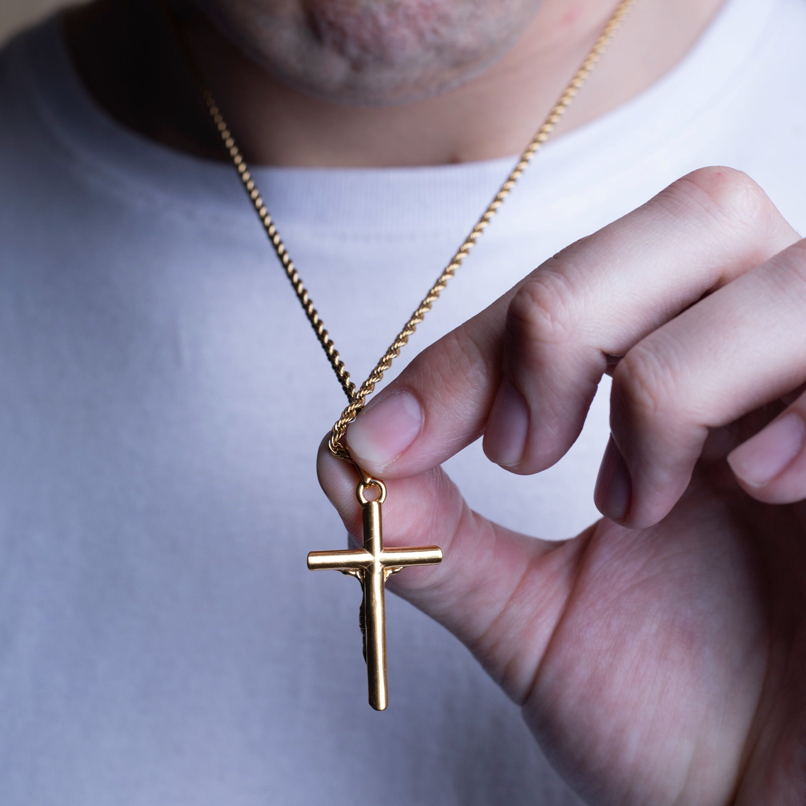 A close-up of a person wearing a white t-shirt and holding a gold cross pendant on a gold chain necklace.
