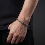 A close-up of a person's wrist wearing a braided gray rope bracelet with a small gold-toned cylindrical bead accent, paired with a dark textured shirt.