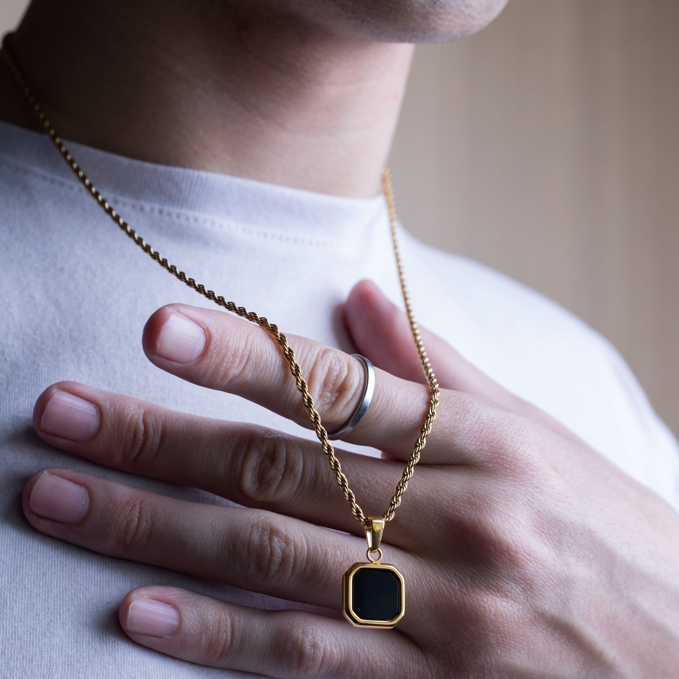 A close-up of a person wearing a gold chain necklace with a square black pendant, resting their hand on their chest.