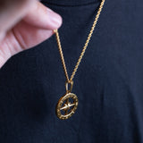 A gold compass pendant necklace worn by a person in a black t-shirt, with a hand holding the chain in the foreground.