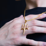 A gold pendant necklace featuring a stylized trident or three-prong symbol, worn on a gold chain and displayed on a person's hand against a dark navy shirt and black background.