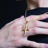 A gold pendant necklace featuring a stylized trident or three-prong symbol, worn on a gold chain and displayed on a person's hand against a dark navy shirt and black background.