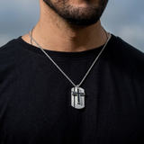 Man wearing a black shirt with a silver necklace featuring a cross pendant against a blurred background