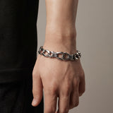 A close-up of a person's wrist wearing a chunky silver chain bracelet with textured details on the links, set against a neutral beige background.