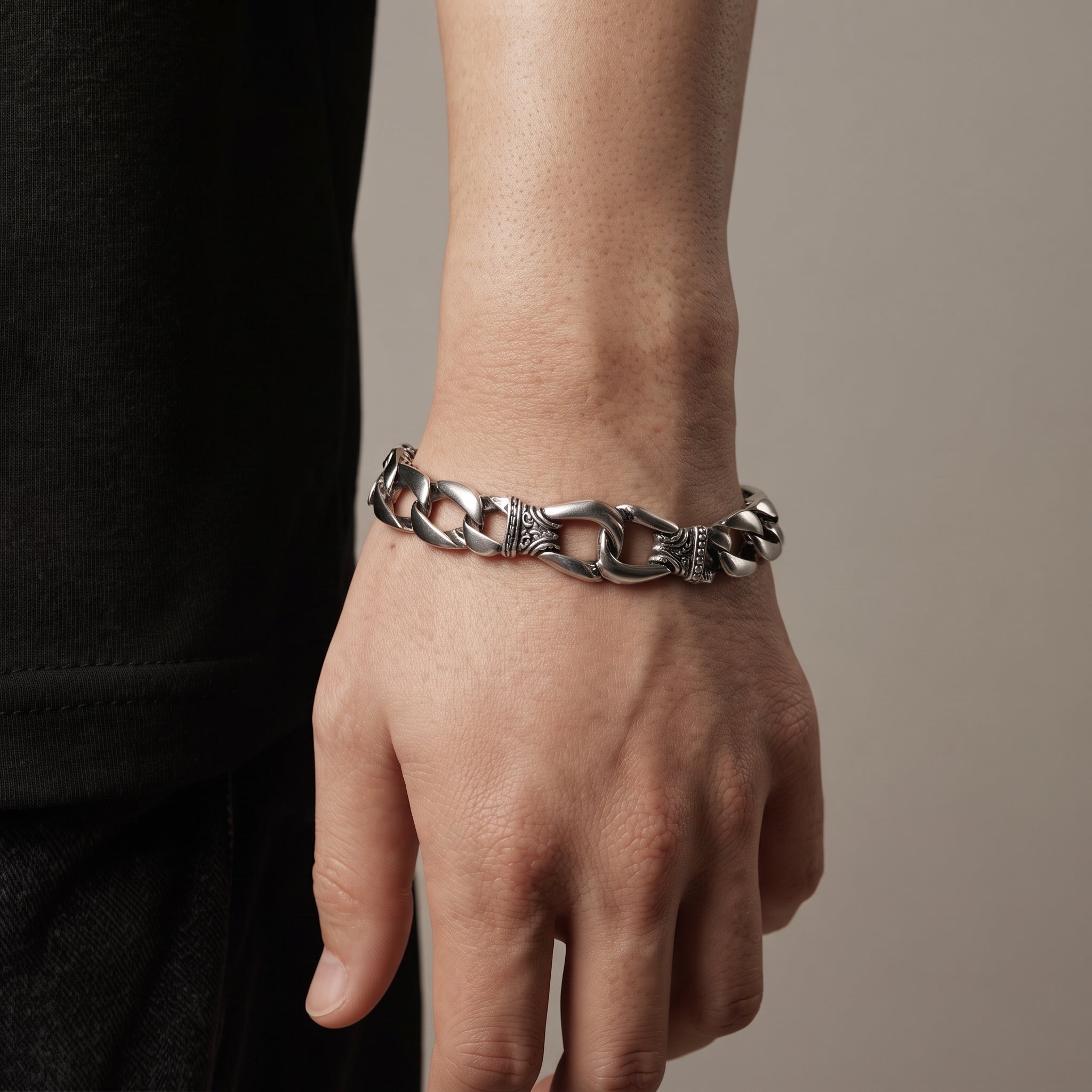 A close-up of a person's wrist wearing a chunky silver chain bracelet with textured details on the links, set against a neutral beige background.