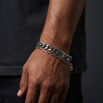 A close-up of a person's wrist wearing a chunky silver-tone chain bracelet with a small round charm, paired with a thinner matching chain bracelet.