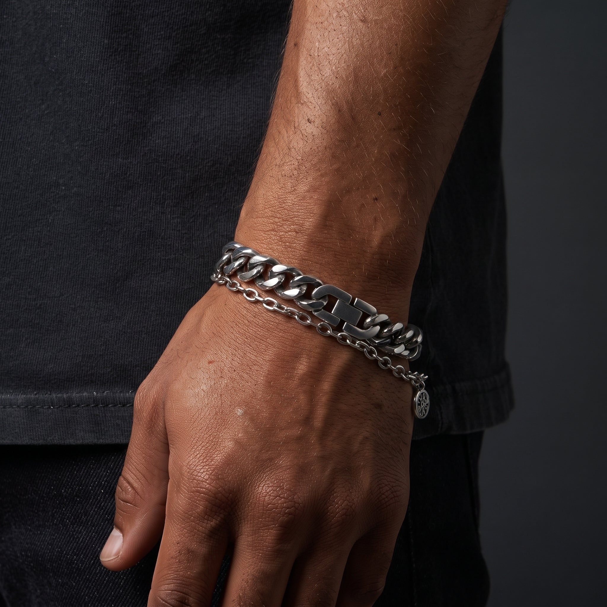 A close-up of a person's wrist wearing a chunky silver-tone chain bracelet with a small round charm, paired with a thinner matching chain bracelet.