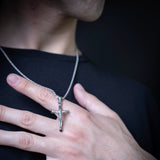 A close-up of a silver crucifix pendant on a chain, worn on a person's finger over a black shirt.