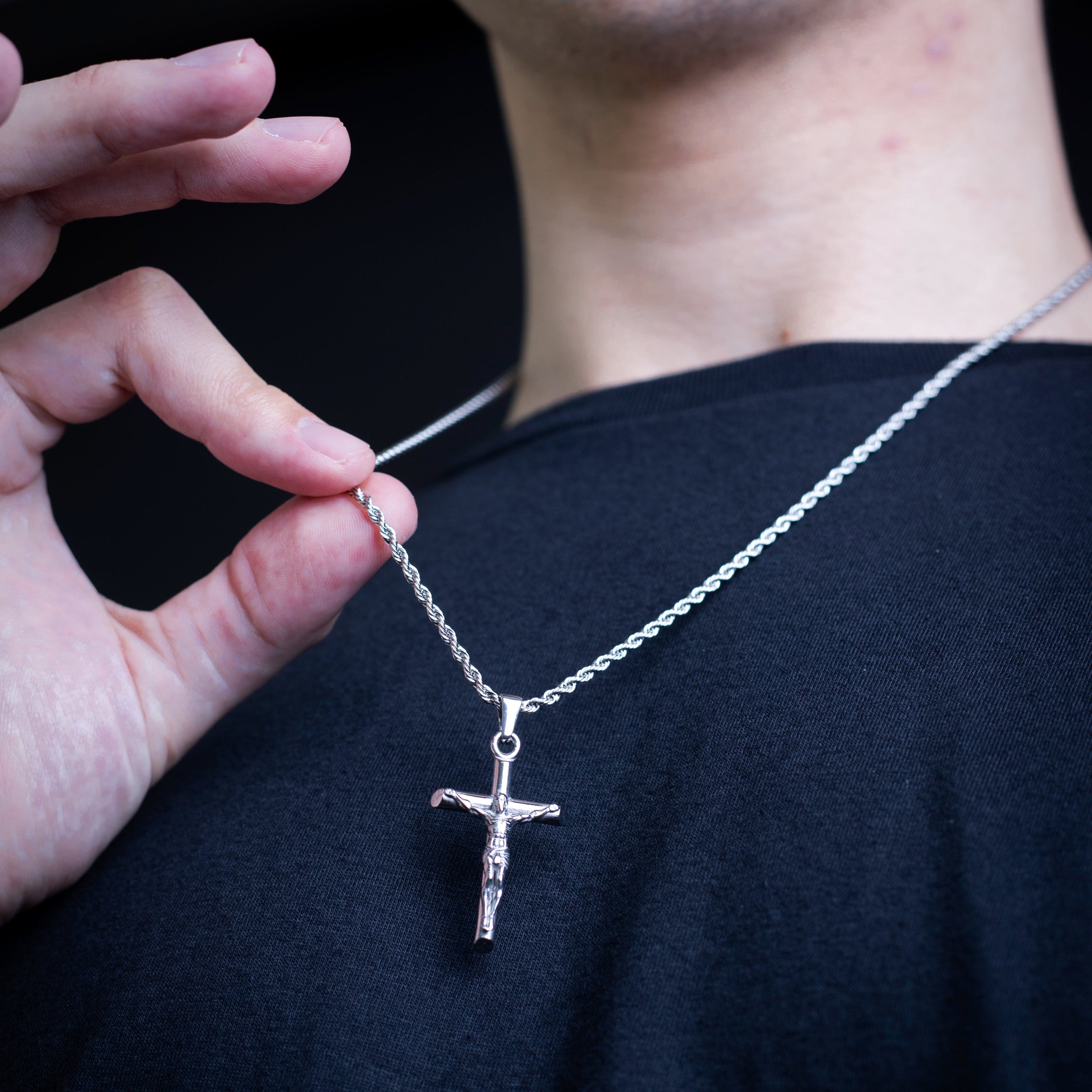 A close-up of a silver crucifix pendant on a chain, held by a person's hand against their black shirt.